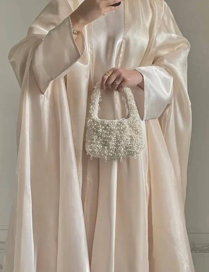 A woman wearing a soft satine finish drape abaya in white, holding a pearl bag