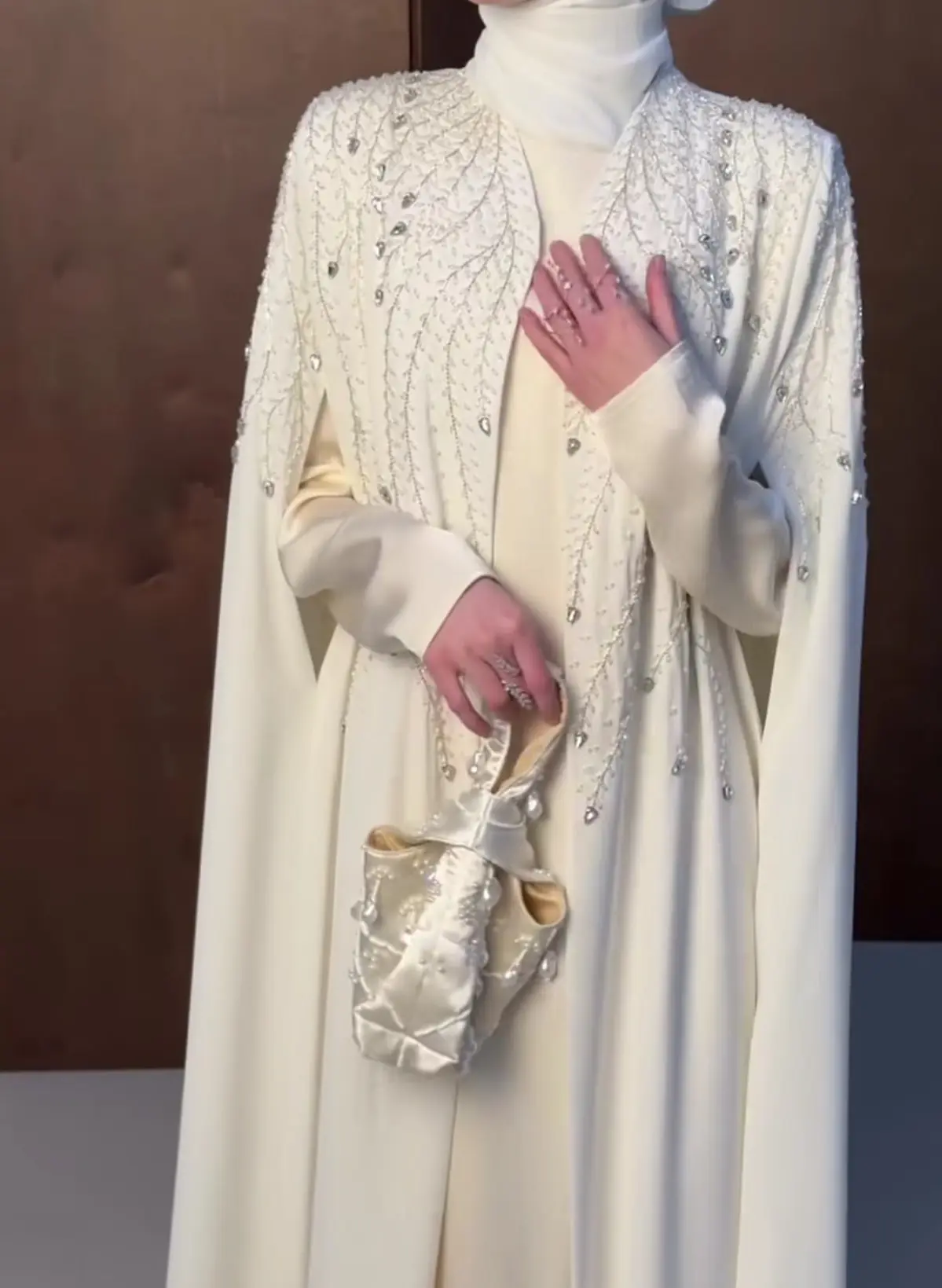  A woman wearing an ivory embellished cap-style abaya, featuring a white hijab and a flowing white dress