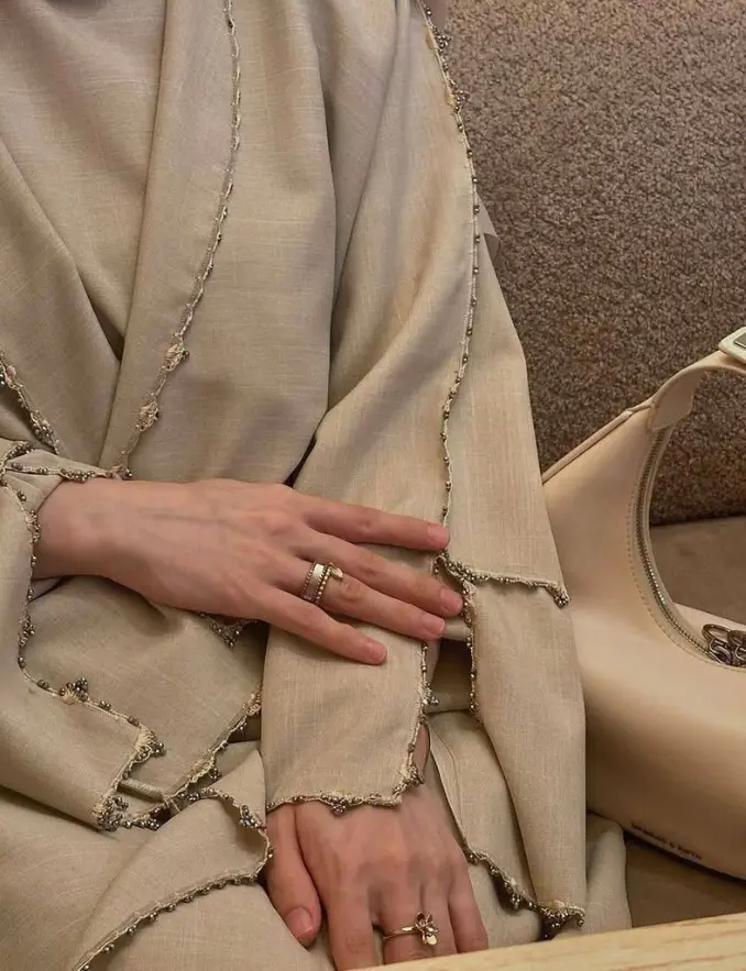 A woman in a beige crystal edge layered abaya sits comfortably on a couch, exuding a relaxed and elegant demeanor