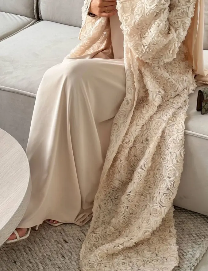 A woman in a beige lace comfort abaya and white dress sits gracefully on a couch, exuding elegance and tranquility