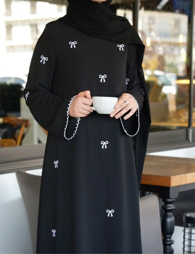 A woman in a black abaya with minimal bow embroidery, holding a cup of coffee, smiling softly