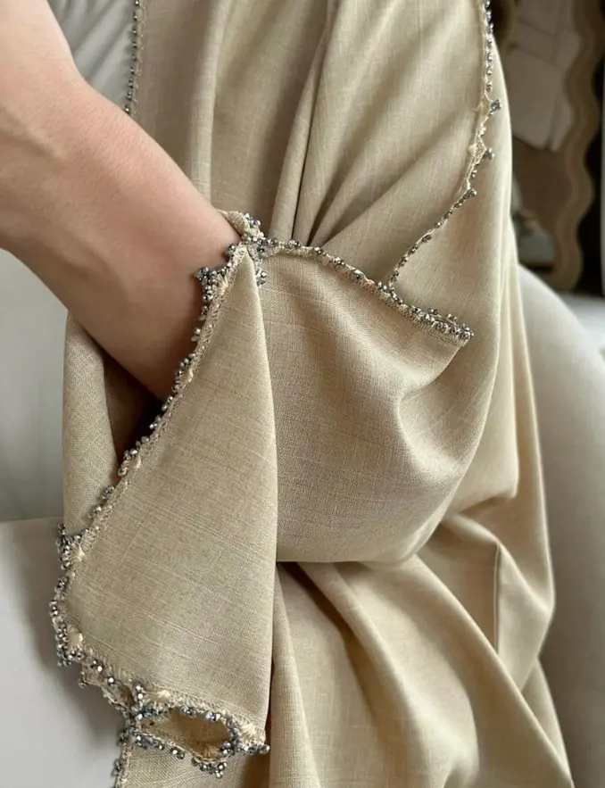 A woman in a beige shawl holds a phone, showcasing the elegant Sand Crystal Trim Sleeve Abaya she is wearing