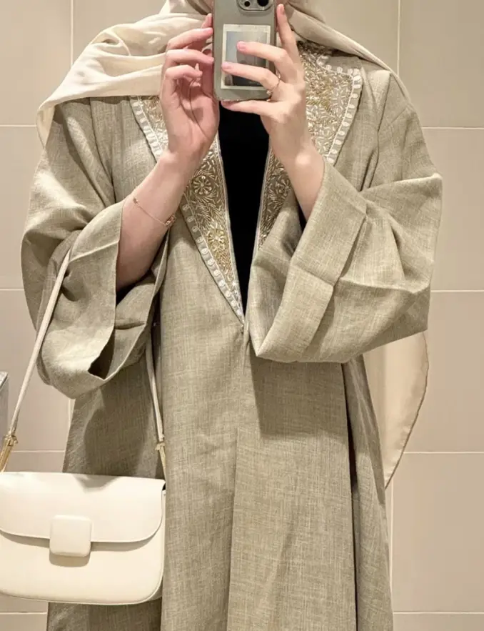 A woman in a hijab wearing a soft taupe everyday abaya takes a selfie in a bathroom mirror